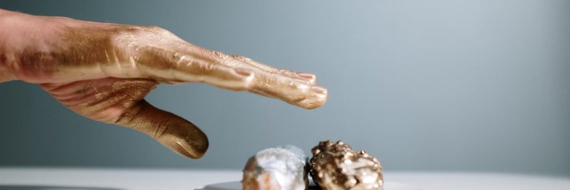 A golden-painted hand reaches towards artfully decorated pastries on a white plate.